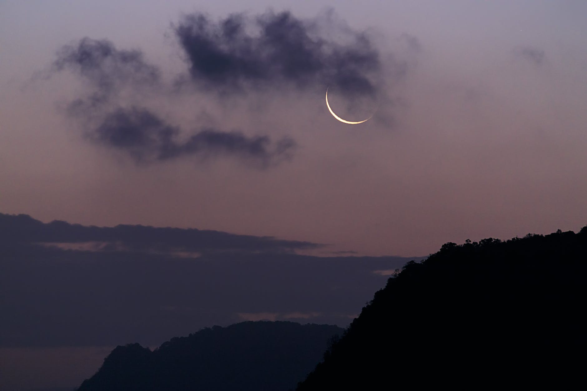 Scene looking over hills. Darkening sky with small crescent moon showing behind a wispy cloud.