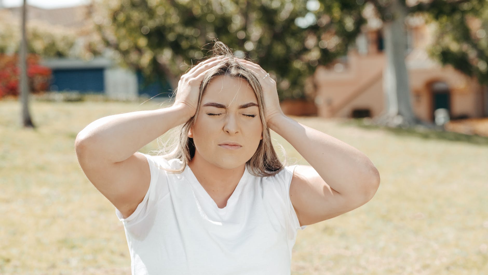 Woman sit outside in a plain white tshirt and her hands on the side of her head as if to quiet her mind